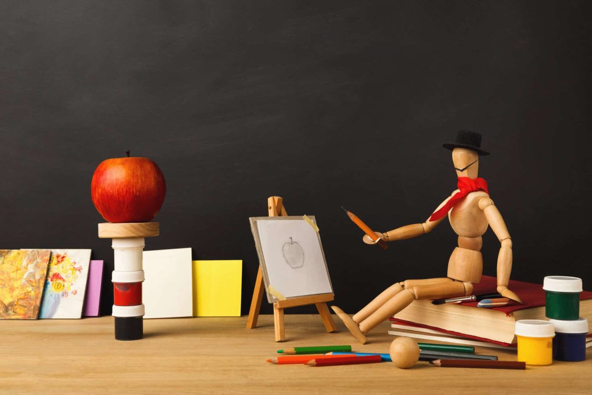 Art background. Wooden marionette artist in hat sitting on old book and drawing an apple against empty classroom blackboard, copy space. Back to school concept